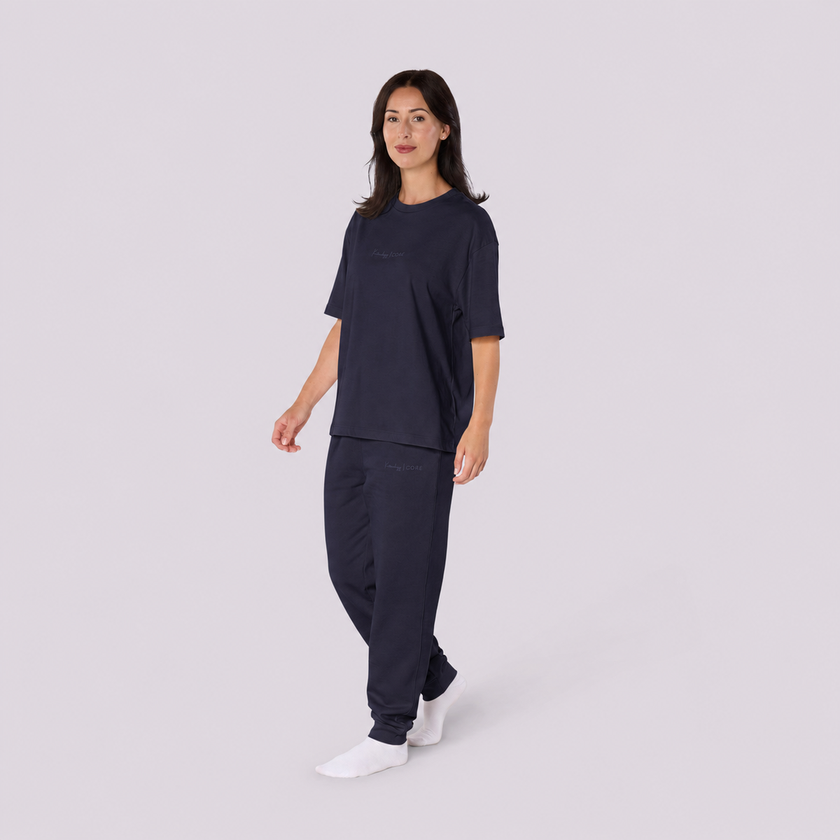 Woman walking in a French navy Kottonology CORE organic cotton T shirt and matching joggers, against a light grey studio background.